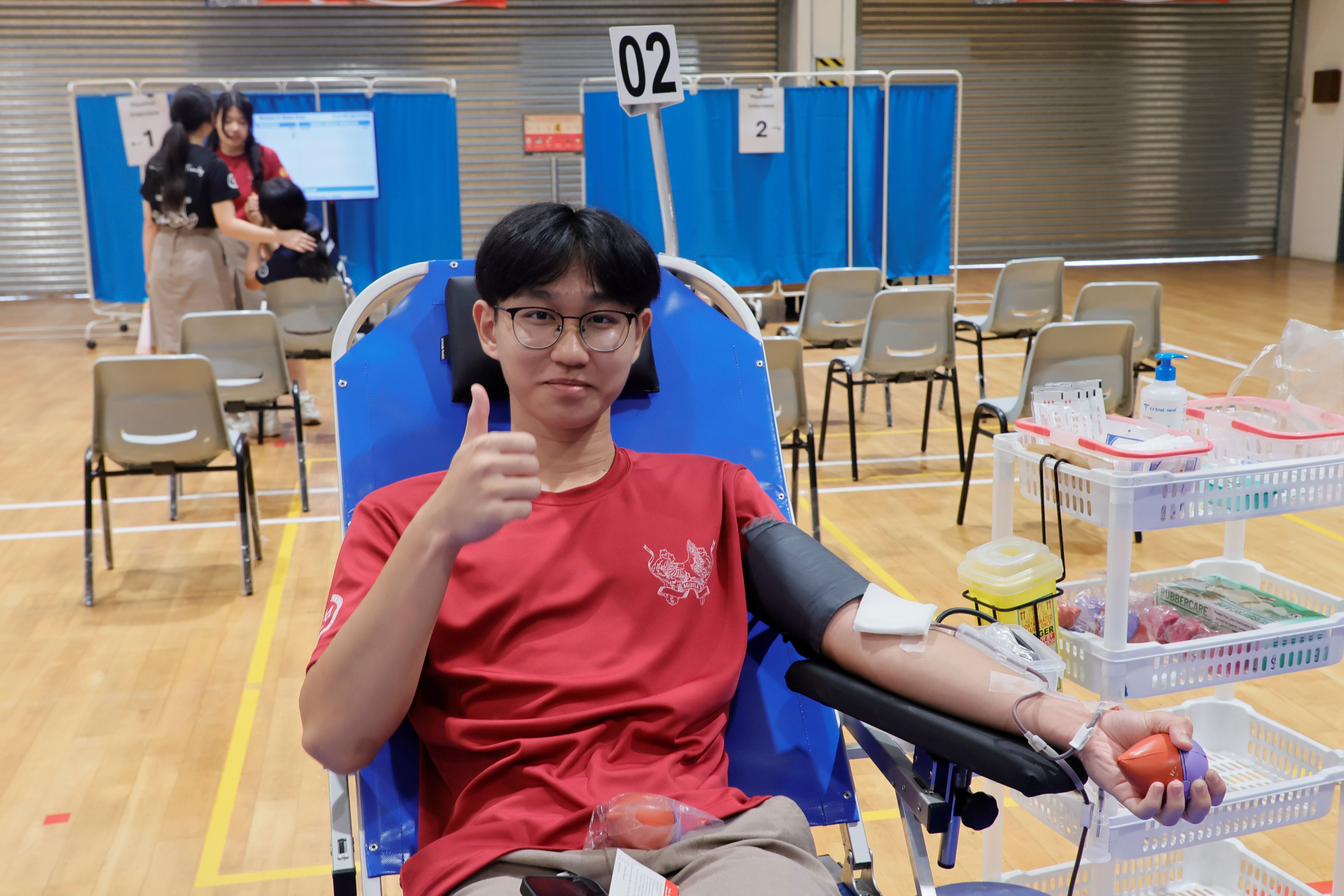 A young male student in a red TMJC t-shirt gives a thumbs-up while donating blood, seated in a donation chair with a blood pressure cuff on his arm. Medical supplies are visible on a trolley beside him.